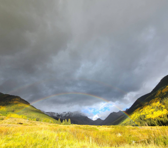Double rainbow over the mountains near Ophir, Colorado, with golden fall aspens illuminated beneath dark storm clouds.